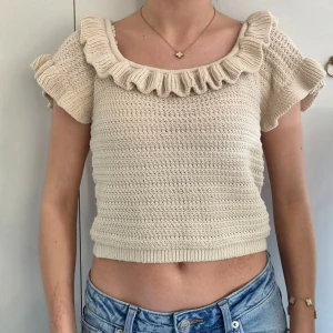 Beige virkad crop top med volang - Söt beige virkad crop top med volangdetaljer längs urringningen och axlarna. Toppen har korta ärmar och kan bäras både på axeln och off-shoulder. Tillverkad i mjukt, stickat material som ger en bohemisk och trendig look.