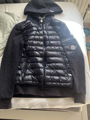 Svart Moncler cardiganjacka med huva - Snygg svart cardiganjacka från Moncler med quiltad front och ärmficka med Moncler-logga. Jackan har huva med dragsko, två dragkedjefickor fram och mjuka ärmar i sweatshirtmaterial. Perfekt för lager på lager och streetwear-stil.