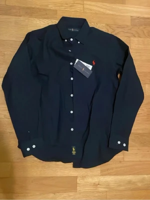 Marinblå skjorta från Ralph Lauren - Klassisk marinblå skjorta från Ralph Lauren med vit knappstängning och röd broderad logga på bröstet. Skjortan har lång ärm och är tillverkad i bomull, perfekt för en stilren look.