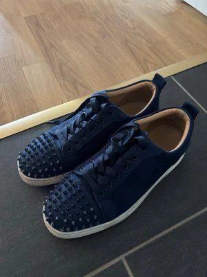Mörkblå sneakers med nitar - Snygga mörkblå sneakers från Christian Louboutin med coola nitar på tån och ovandelen. Skorna har en låg profil, rund tå och är gjorda i mocka med detaljer i läder. Vita sulor och matchande blå skosnören ger en stilren look. Skorna är inte vid jätte bra skick vi spikarna och har tappat 4-5 st
