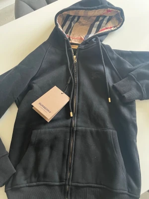 Svart hoodie med dragkedja Burberry - Snygg svart hoodie från Burberry med dragkedja framtill och klassiskt rutigt foder i huvan. Hoodien har ribbade muddar, fickor fram och guldfärgade detaljer på snörena. Perfekt för dig som vill ha en stilren och exklusiv look.