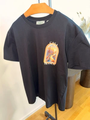 Svart t-shirt från Casablanca - Snygg svart t-shirt från Casablanca med tryck framtill som visar en solnedgång och texten 'Casa Blanca'. Klassisk rund hals och korta ärmar. Perfekt för dig som gillar streetwear och vill ha något unikt i garderoben.