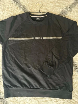 Svart sweatshirt från BOSS - Svart sweatshirt från BOSS med rund halsringning och diskret logga framtill. Tröjan har en silver horisontell linje över bröstet och ribbade muddar vid ärmslut och nederkant. Perfekt för en avslappnad och stilren look. Storlek M passar S bättre passar inte mig tyvärr…