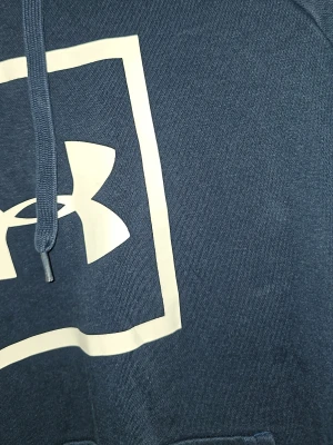 Mörkblå hoodie från Under Armour - Snygg mörkblå hoodie från Under Armour med stor vit logga i fyrkant på bröstet. Klassisk känguruficka framtill, dragsko i huvan och ribbade muddar. Perfekt för dig som gillar sportig och avslappnad stil. Är öppen för prisförslag vid snabb försäljning.