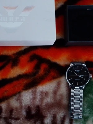 Herr klocka från Nibosi - Stilren klocka från Nibosi med svart urtavla och datumvisning. Armbandet är i silverfärgad metall med klassisk länkdesign. Kommer med originalbox och garanti.