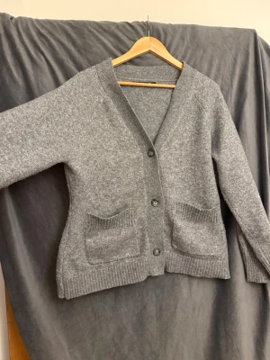 Grå stickad kofta med fickor - Mysig grå stickad kofta med v-ringning och tre knappar framtill. Koftan har två rymliga fickor framtill och ribbade muddar vid ärmslut och nederkant. Perfekt för lager på lager och chill dagar.