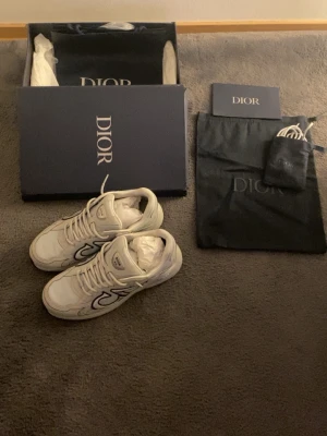 Vita Dior B30 sneakers med reflexdetaljer - Säljer ett par vita Dior B30 sneakers med grå och silvriga reflexdetaljer på sidan. Skorna har en chunky sula och tydlig Dior-logga. Tillverkade i en mix av mesh och läder för en modern look. Levereras med originalkartong, dustbag och extra skosnören.