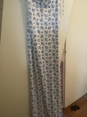 Vit maxiklänning med blått blommönster - Lång maxiklänning i vitt med blått blommönster och knytning vid bysten. Klänningen har rak passform och tunna axelband. Perfekt för sommarens varma dagar och ger en romantisk look.