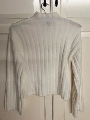 Vit ribbstickad polotröja - Säljer en vit ribbstickad polotröja med hög hals och långa ärmar. Tröjan är mjuk och har en relaxed fit som passar perfekt till kalla vintrar. Håller sig varm och skön. Materialet känns fluffigt och mysigt.