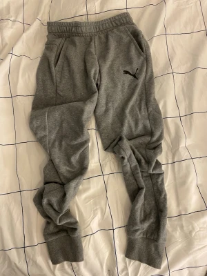 Gråa mjukisbyxor från Puma - Säljer ett par gråa mjukisbyxor från Puma med resår i midjan och muddar vid bensluten. Byxorna har två sidofickor och en svart Puma-logga på vänster lår. Perfekta för chill dagar eller träning.