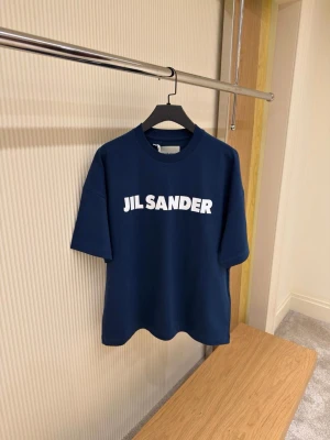 Blå Jil Sander t-shirt med tryck - Snygg mörkblå t-shirt från Jil Sander med stort vitt logotryck på bröstet. Klassisk rund hals och korta ärmar. Tillverkad i mjuk bomull för en skön känsla och clean look. Perfekt för dig som gillar minimalistisk och stilren design.