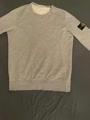 Grå sweatshirt från Stone Island - Snygg grå sweatshirt från Stone Island med klassisk rund hals och ribbade muddar. Tröjan har den ikoniska svarta patchen med gult Stone Island-märke på vänster ärm. Perfekt för en avslappnad och trendig stil.