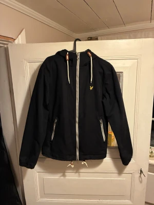 Svart jacka Lyle & Scott - Snygg svart jacka från Lyle & Scott med huva. Perfekt för våren och enkel att matcha med olika outfits.