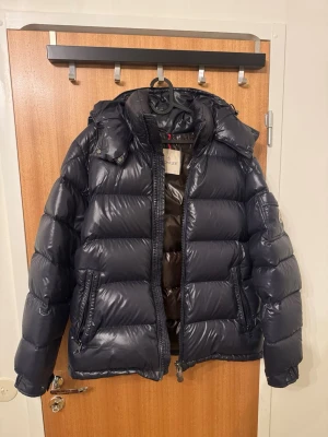 Moncler Maya strl 5 - Säljer en svart, glansig dunjacka från Moncler med avtagbar huva och två stora fickor framtill. Jackan har kraftiga quiltade sektioner och dragkedja. Perfekt för kalla vinterdagar och riktigt snygg att bära till jeans eller joggers.