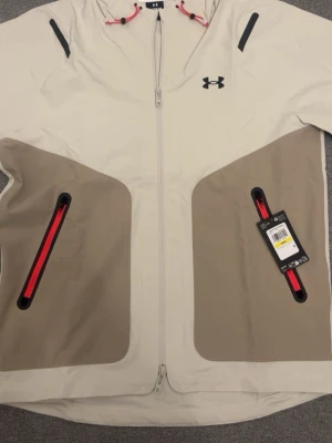 Beige vindjacka från Under Armour - Stilren beige vindjacka från Under Armour med kontrasterande ljusbruna partier och svarta detaljer. Jackan har hel dragkedja, två fickor med röda dragkedjor och justerbar huva med röda snören. Perfekt för blåsiga dagar och sportiga outfits.