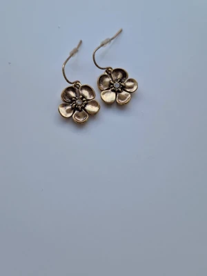 Blommiga örhängen i guldton Ikita från Paris y2k - Snygga örhängen i form av blommor med fem kronblad i guldtonad metall. Hängande modell med krok, detaljerad mittdel och lätt antik finish. Perfekta för att ge din stil en unik touch. Kommer från Ikita Paris. Inköpta början av 2000-talet. 1.5 cm 