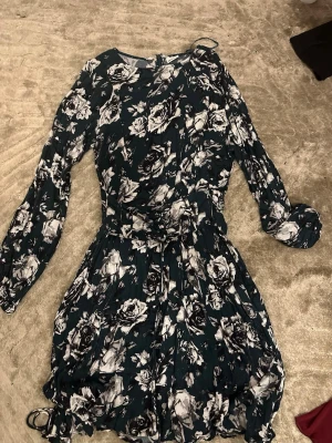 Mörkgrön blommig kortklänning med snörning - Supersnygg kortklänning i mörkgrönt med vitt och svart blommönster. Klänningen har långa ärmar och cool snörningsdetalj i midjan. Priset kan dikteras 