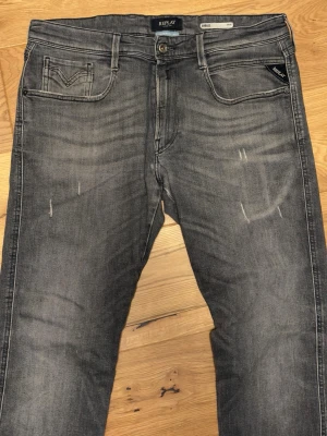 Replay Anbass - Snygga grå slim fit jeans från Replay med slitna detaljer och klassisk femficksdesign. Jeansen har en lätt tvättad look, svarta sömmar på bakfickorna och metallknapp framtill. Perfekta för en avslappnad och trendig stil.
