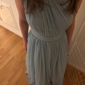 Ljusblå balklänning - Säljer en elegant ljusblå långklänning med omlottdetalj och markerad midja. Klänningen har ett svepande fall och är ärmlös med en axelbandslös look. Perfekt för speciella tillfällen där du vill känna dig extra fin.