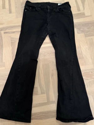 True Religion Svarta utsvängda jeans - Säljer ett par svarta jeans med utsvängda ben och klassisk femficksdesign. Jeansen har låg midja och stängs med knapp och dragkedja. Perfekta för dig som gillar en retroinspirerad look.
