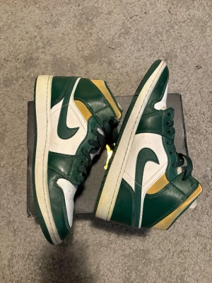 Nike Air Jordan 1 Mid grön/vit/gul - Säljer ett par Nike Air Jordan 1 Mid sneakers i grönt, vitt och gult. Skorna har klassisk hög siluett, gröna snören och detaljer i läder. Ikonisk Swoosh på sidan och Air Jordan-logga vid ankeln. Perfekta för dig som gillar streetwear och retrostil eller gillar att spara på skor.