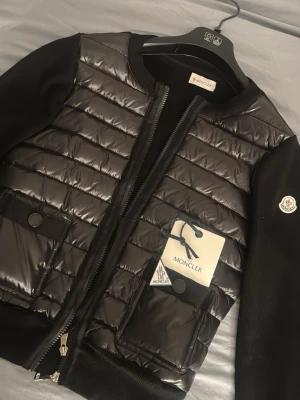 Svart pufferjacka från Moncler - Snygg svart pufferjacka från Moncler med glansig quiltad front, två stora fickor med knappstängning och Moncler-logga på ärmen. Jackan har ribbade muddar och dragkedja framtill. Perfekt för kalla vinterdagar och riktigt stilren look.