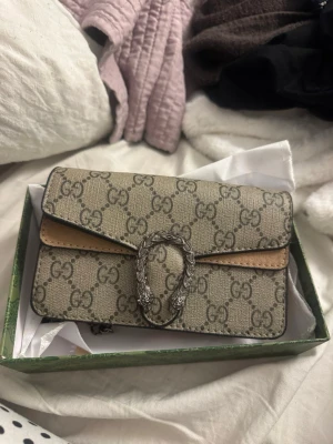 Gucci Dionysus GG Supreme handväska - Säljer en Gucci Dionysus handväska i beige GG Supreme canvas med bruna detaljer och silverfärgat spänne med ormmotiv. Klassisk rektangulär form och ikoniskt GG-mönster över hela väskan. Perfekt för dig som vill ha en lyxig och trendig accessoar.