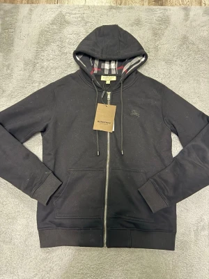 Svart hoodie med dragkedja från Burberry - Svart hoodie från Burberry med dragkedja framtill och klassiskt rutigt foder i huvan. Stora fickor framtill, ribbade muddar och justerbar huva med snören. Perfekt för en stilren och avslappnad look.