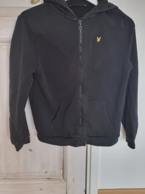Svart hoodie från Lyle & Scott - Svart hoodie med dragkedja från Lyle & Scott. Klassisk gul logga på bröstet, huva med snörning och känguruficka framtill. Tillverkad i mjukt bomullsmaterial, perfekt för en avslappnad stil.