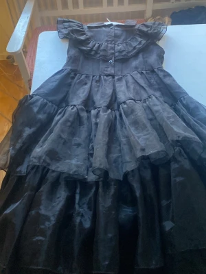 Svart wednesday klänning  - Säljer en svart wednesday klänning med flera lager volanger och offshoulder-design. Klänningen har knappar framtill och är tillverkad i ett tunt, glansigt material som ger en elegant look. Perfekt för dig som vill sticka ut med en dramatisk och trendig stil.