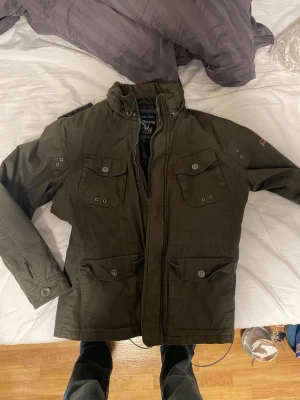 Olivgrön vindjacka från Crafted Goods - Snygg olivgrön vindjacka från Crafted Goods med fyra framfickor med lock och knapp, hög krage och dragkedja. Jackan har en klassisk militärinspirerad look och är tillverkad i ett slitstarkt material med svart innerfoder. Jackan är för stor och jag är ca 186 70 kg✌🏼