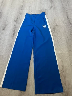 Blåa mjukisbyxor med vita ränder - Snygga blå mjukisbyxor med raka ben och vita ränder längs sidorna. Byxorna har broderad vit logga 'ISETTEPA' och 'TB' på ena benet samt fickor framtill. Perfekta för en sportig och avslappnad stil.