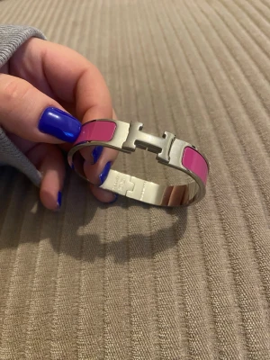 H rosa Clic H armband - Snyggt armband från H i modellen Clic H. Armbandet är i silverfärgad metall med rosa emaljdetaljer och har det ikoniska H-spännet framtill. Perfekt statement piece för att lyfta din outfit med en pop av färg.