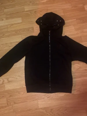 C.P. Company hoodie - Säljer en svart hoodie från C.P. Company med hel dragkedja och ikoniska glasögon i huvan. Hoodien har ribbade muddar och en cool streetwear-look. Perfekt för dig som vill sticka ut med en unik detalj på huvan. Bra gick använt ett par gånger men skickas med fint skick som helt ny. Priset går att diskuteras vid snabb affär.