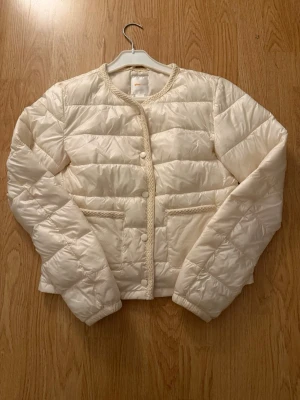 Krämvit jacka från Gina Tricot - Snygg krämvit jacka från Gina Tricot med rund halsringning och dekorativa stickade detaljer längs kanterna. Västen har tryckknappar framtill och två fickor. Perfekt för lager på lager och ger en clean look till din outfit.