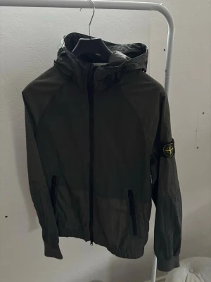 Mörkgrön vindjacka från Stone Island - Snygg mörkgrön vindjacka från Stone Island med huva och dragkedja framtill. Jackan har två fickor med dragkedja och den klassiska Stone Island-loggan på vänster ärm. Tillverkad i lätt, vindtätt material som passar perfekt för blåsiga dagar.
