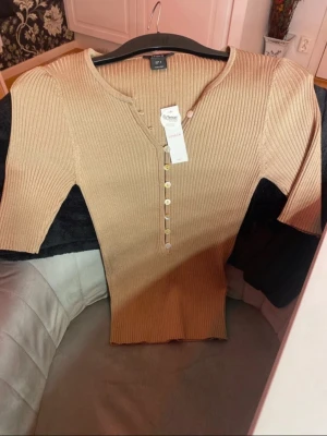 Beige ribbstickad topp från Lindex - Trendig beige/aprikos ribbstickad topp från Lindex med korta ärmar och snygg knappdetalj framtill. Tillverkad i mjukt Tencel-material som känns skönt mot huden. Perfekt för dig som gillar en enkel men stilren look.