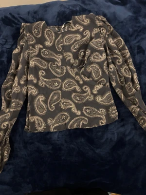 Svart paisleymönstrad blus från Zara - Snygg svart blus från Zara med beige paisleymönster. Blusen har långa ärmar och rund halsringning. Tillverkad i ett tunt, mjukt tyg som ger en skön känsla och ett elegant fall. Perfekt för dig som gillar trendiga mönster och stilrena detaljer.