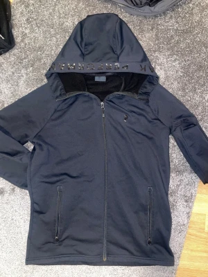 Svart hoodie från Peak Performance - Snygg svart hoodie från Peak Performance med dragkedja framtill och två fickor med dragkedja. Huva med tryckt text och mjukt fleecefoder på insidan. Perfekt för dig som gillar sportig och clean stil.