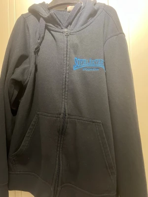 Svart sofialäktaren zip up - Svart hoodie från Clique med dragkedja och huva. Broderad text 'Sofialäktaren Stockholm' i blått på bröstet. Stora fickor framtill och justerbar dragsko i huvan. Perfekt för dig som gillar enkel och stilren streetwear. Sällsynt zip up från 2021.