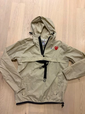 Beige vindjacka från Comme des Garçons - Snygg beige vindjacka från Comme des Garçons PLAY med ikoniskt rött hjärta på bröstet. Jackan har huva, halv dragkedja framtill, justerbar midja och svarta detaljer. Tillverkad i lätt och tunt material, perfekt för blåsiga dagar.