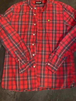 Röd rutig skjorta från Lyle & Scott - Klassisk röd rutig skjorta från Lyle & Scott med lång ärm och vita knappar. Skjortan har en gul logga broderad på bröstet. Perfekt för en avslappnad men snygg stil.