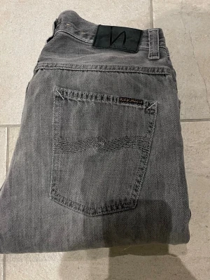 Grå raw denim Nudie Jeans - Säljer ett par grå jeans från nudie jeans i modellen loose Leif. Jeansen har en snygg tvättad look och detaljerad söm på bakfickan. Tillverkade i raw denim, perfekta för en avslappnad stil. Jeans har tvyörr fått en heel bite 30/34