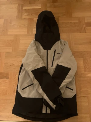 Svart och beige skidjacka från Everest - Snygg skidjacka från Everest i svart och beige med huva och dragkedja framtill. Jackan har flera fickor med dragkedja och en sportig design med kontrastpaneler. Perfekt för blåsiga dagar och outdoor-aktiviteter.