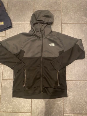 Svart hoodie från The North Face - Snygg svart hoodie från The North Face med dragkedja framtill och två fickor med dragkedja. Hoodien har en tvåfärgad design med mörkgrå upptill och svart nedtill samt en vit logga på bröstet. Perfekt för en avslappnad streetstyle-look.