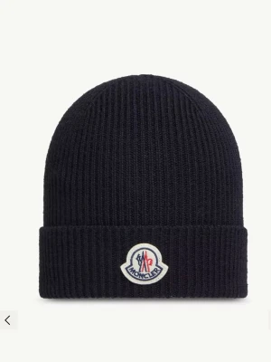 Svart Moncler ribbstickad mössa - Svart ribbstickad mössa från Moncler med uppvikt kant och ikonisk Moncler-logga framtill. Tillverkad i mjukt material som ger både värme och stil. Enkel, clean design som funkar till det mesta och ger en exklusiv känsla.