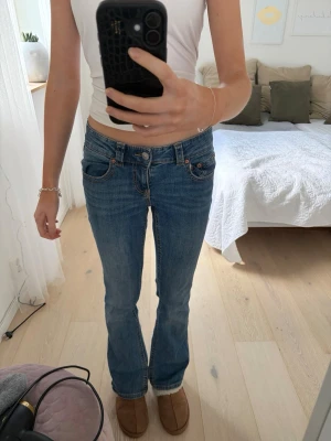 Blå bootcut jeans med fickdetaljer - Säljer ett par blå bootcut jeans med låg midja och snygga fickdetaljer bak med knappar. Jeansen har klassisk femficksmodell och är i ett mjukt denimtyg. Perfekta för en trendig look.