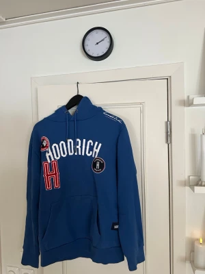 Blå hoodie från Hoodrich - Snygg blå hoodie från Hoodrich med vita och röda detaljer samt broderade patches på bröstet. Stor ficka framtill och justerbar huva med dragsko. Hoodien har en avslappnad passform och är tillverkad i mjukt material, perfekt för en streetwear-look.