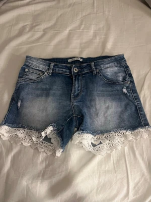 Blå jeansshorts med spetskant - Säljer ett par blå denimshorts från Lace & Beads med snygga slitningar och vit spetskant längst ner. Klassisk femficksmodell med knapp och dragkedja framtill. Perfekta för sommaren och ger en söt touch till din outfit!