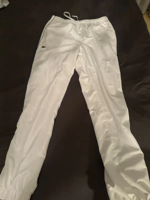 Vita träningsbyxor från Lacoste Sport - Säljer ett par vita träningsbyxor från Lacoste Sport med elastisk midja och snörning. Byxorna har den klassiska gröna krokodilloggan på vänster sida och är tillverkade i ett lätt, syntetiskt material. Regular fit och perfekta för sportiga dagar.